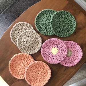 May include: Six handmade crochet coasters in beige, green, pink, and peach. Two pink coasters feature a yellow centre. The coasters are displayed on a dark wooden surface.
