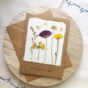 Peut inclure: Une carte de vœux rectangulaire avec une bordure en papier kraft et un panneau central blanc orné de fleurs séchées pressées dans des tons de violet et de jaune. Le texte Birthday Wishes est imprimé en écriture noire en bas de la carte.