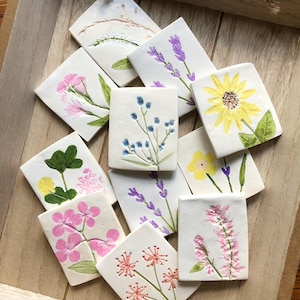 May include: A selection of square, white ceramic tiles, each hand-painted with a different floral design. The flowers include lavender, sunflowers, and pink blossoms. The tiles are arranged in a wooden tray, showcasing the variety of botanical art.