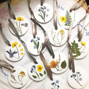 May include: A collection of round white ceramic ornaments, each featuring a unique pressed flower or fern specimen. Every ornament is suspended by a thin, smooth taupe-coloured ribbon, showcasing botanical details in yellow, blue, and green hues.