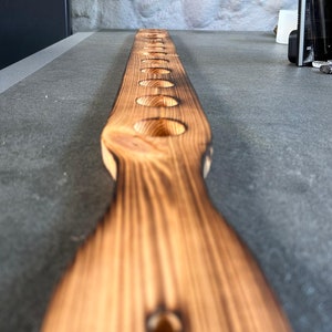 Pu&ograve; includere: Un lungo asse di legno di colore arancione bruciato con una serie di fori circolari equidistanti praticati. L'asse &egrave; posto piatto su una superficie grigia.