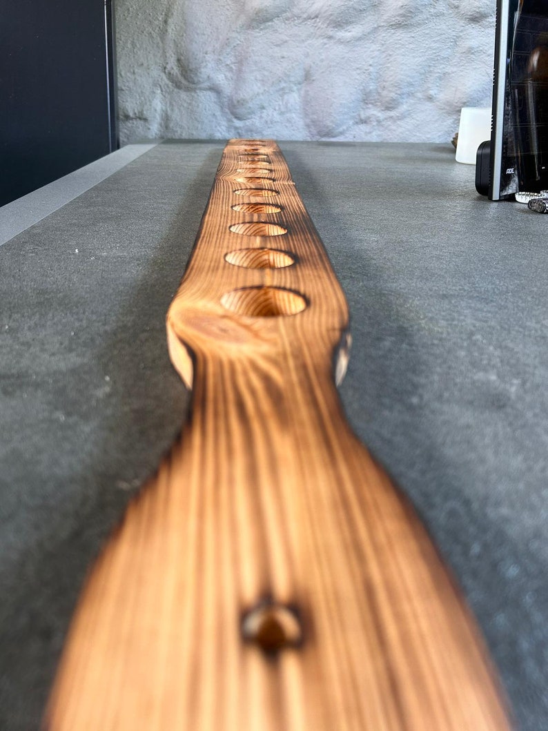 Pu&ograve; includere: Un lungo asse di legno di colore arancione bruciato con una serie di fori circolari equidistanti praticati. L'asse &egrave; posto piatto su una superficie grigia.