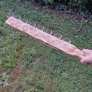Op de afbeelding: Een houten serveerplank met acht kleine borrelglaasjes. De plank is gemaakt van lichtgekleurd hout en heeft een natuurlijke, rustieke uitstraling. De glazen zijn helder en leeg, klaar om gevuld te worden. De plank wordt boven een grasveld gehouden.