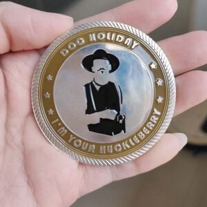 May include: A silver and gold-toned coin with the text "DOG HOLIDAY I'M YOUR HUCKLEBERRY" around the edge. The coin features a silhouette of a man in a hat and suspenders. The coin is held in a hand.