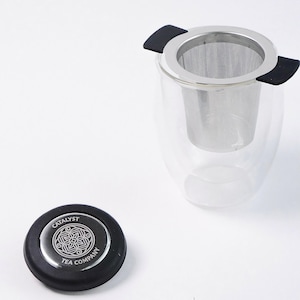 May include: A clear glass tea infuser with a stainless steel mesh filter and black silicone handles. The infuser is sitting on a white surface. A black lid with the text "Catalyst Tea Company" is also shown.
