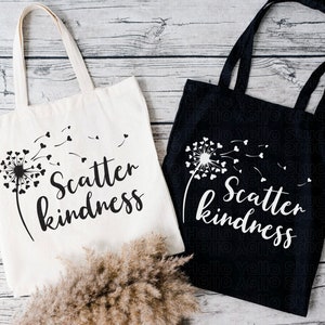 May include: Two tote bags with the text "Scatter kindness" printed on them. One bag is white with black text and a black dandelion design. The other bag is black with white text and a white dandelion design.