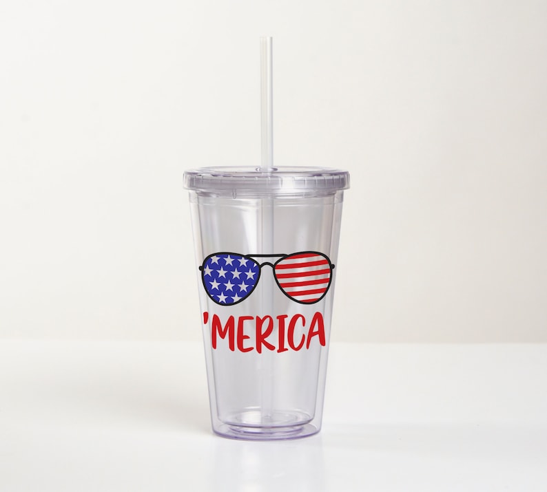 May include: Clear plastic tumbler with a straw, featuring a red, white, and blue American flag design with sunglasses and the text 'MERICA'.