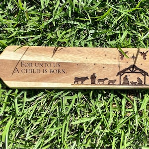 May include: A wooden Christmas ornament with the text "For unto us a child is born." and a silhouette of the nativity scene.