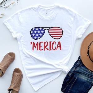 May include: White t-shirt with a graphic of sunglasses with the American flag design and the text 'MERICA' in red.