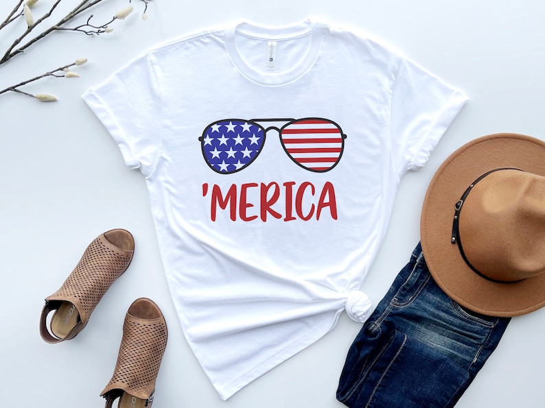 May include: White t-shirt with a graphic of sunglasses with the American flag design and the text 'MERICA' in red.