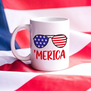 May include: White ceramic mug with a red, white, and blue design of sunglasses with the word 'MERICA' printed below. The mug is sitting on a red, white, and blue American flag.