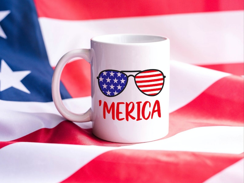 May include: White ceramic mug with a red, white, and blue design of sunglasses with the word 'MERICA' printed below. The mug is sitting on a red, white, and blue American flag.