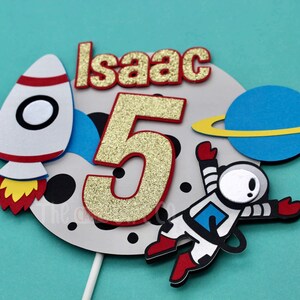 Puede incluir: Un adorno para pastel con un cohete, un astronauta, un planeta y el nombre "Isaac" con el n&uacute;mero 5 en purpurina dorada. El adorno es perfecto para una fiesta de cumplea&ntilde;os con tem&aacute;tica espacial.