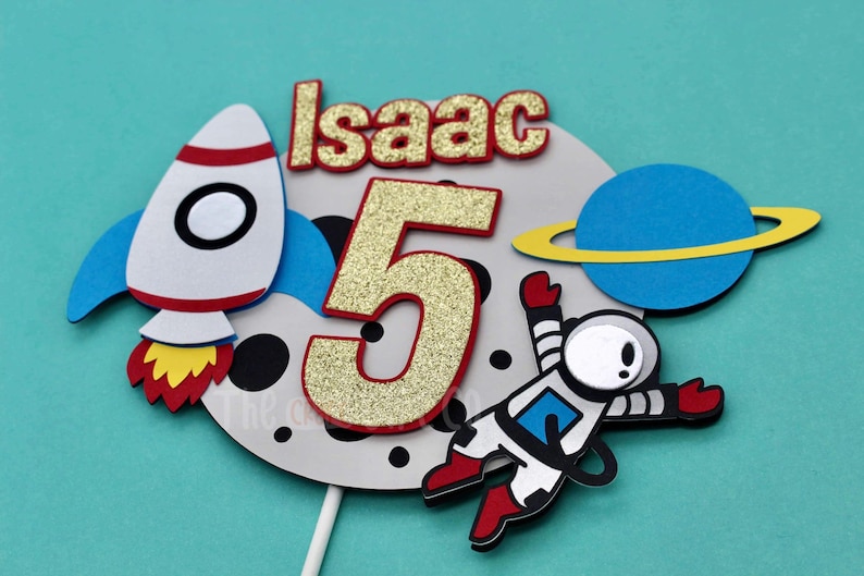 Puede incluir: Un adorno para pastel con un cohete, un astronauta, un planeta y el nombre "Isaac" con el n&uacute;mero 5 en purpurina dorada. El adorno es perfecto para una fiesta de cumplea&ntilde;os con tem&aacute;tica espacial.