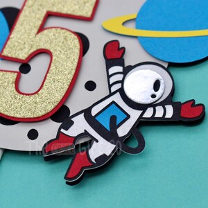 Puede incluir: Un adorno para pastel con un astronauta blanco con botas y guantes rojos, un n&uacute;mero "5" de purpurina dorada, un planeta azul con un anillo amarillo y lunares negros sobre un fondo gris.
