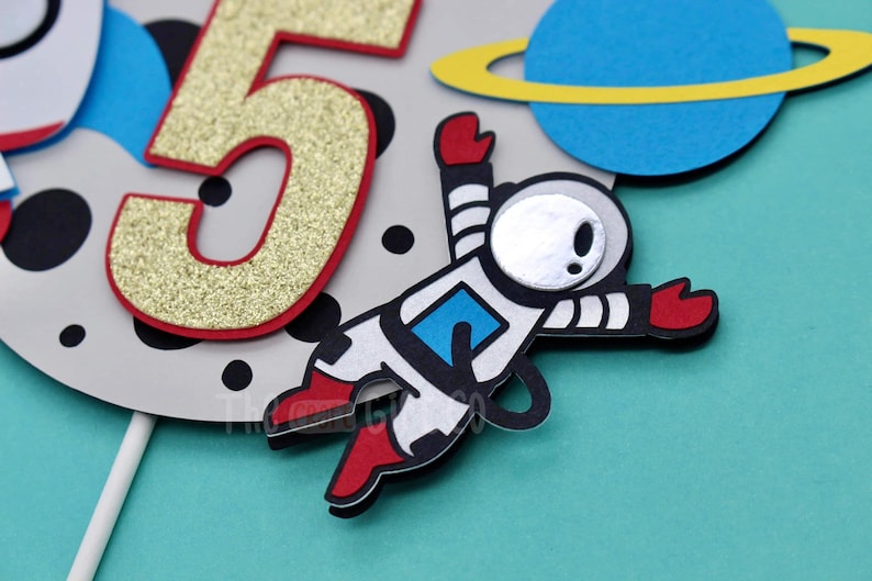 Puede incluir: Un adorno para pastel con un astronauta blanco con botas y guantes rojos, un n&uacute;mero "5" de purpurina dorada, un planeta azul con un anillo amarillo y lunares negros sobre un fondo gris.