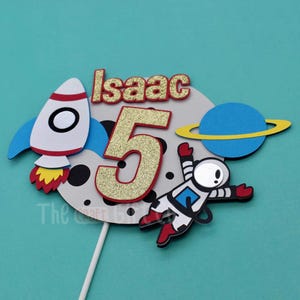 Puede incluir: Un adorno para pastel con un cohete, un astronauta, un planeta y el n&uacute;mero 5 con brillantina. La luna es gris con puntos negros y el texto "Isaac" est&aacute; en la parte superior.