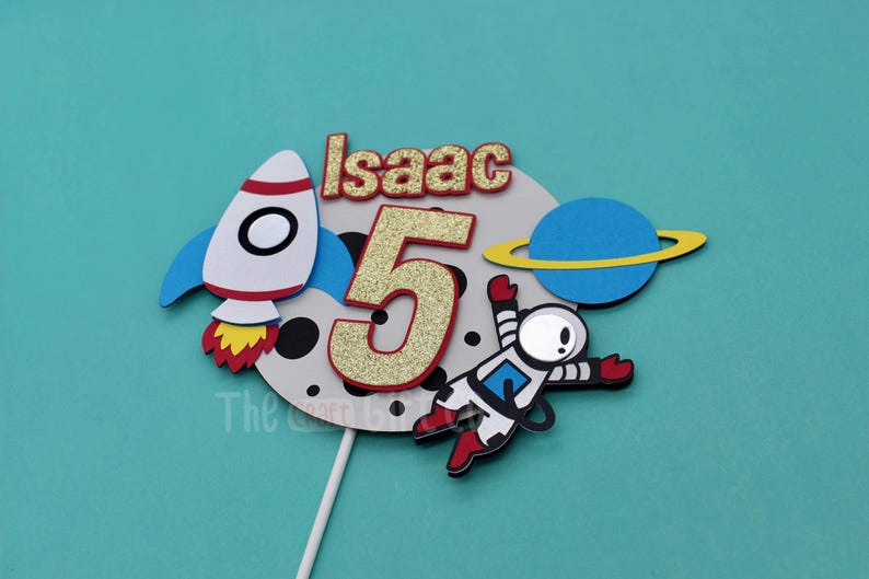 Puede incluir: Un adorno para pastel con un cohete, un astronauta, un planeta y el n&uacute;mero 5 con brillantina. La luna es gris con puntos negros y el texto "Isaac" est&aacute; en la parte superior.