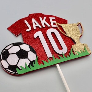 May include: Red and white football shirt cake topper with the name "Jake" and the number "10" on it. A gold trophy and a black and white football are also included. The topper is on a green grass base.