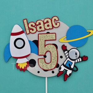 Puede incluir: Un adorno para pastel con un cohete, un astronauta, un planeta y el nombre "Isaac" con un n&uacute;mero 5 brillante. El adorno es perfecto para una fiesta de cumplea&ntilde;os con tem&aacute;tica espacial.