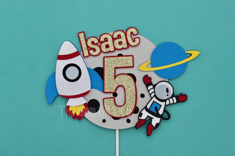Puede incluir: Un adorno para pastel con un cohete, un astronauta, un planeta y el nombre "Isaac" con un n&uacute;mero 5 brillante. El adorno es perfecto para una fiesta de cumplea&ntilde;os con tem&aacute;tica espacial.