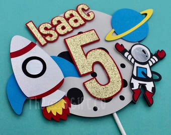 May include: A cake topper featuring a rocket, astronaut, planet, and the number 5 with glitter. The topper also includes the name "Isaac" in gold letters.