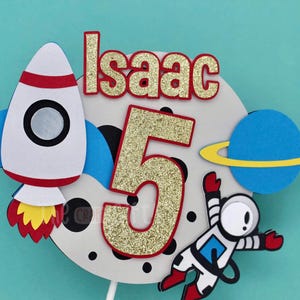 Puede incluir: Un adorno para pastel blanco con un cohete, un astronauta, un planeta y el n&uacute;mero 5 en purpurina dorada. El texto "Isaac" est&aacute; escrito en letras rojas.
