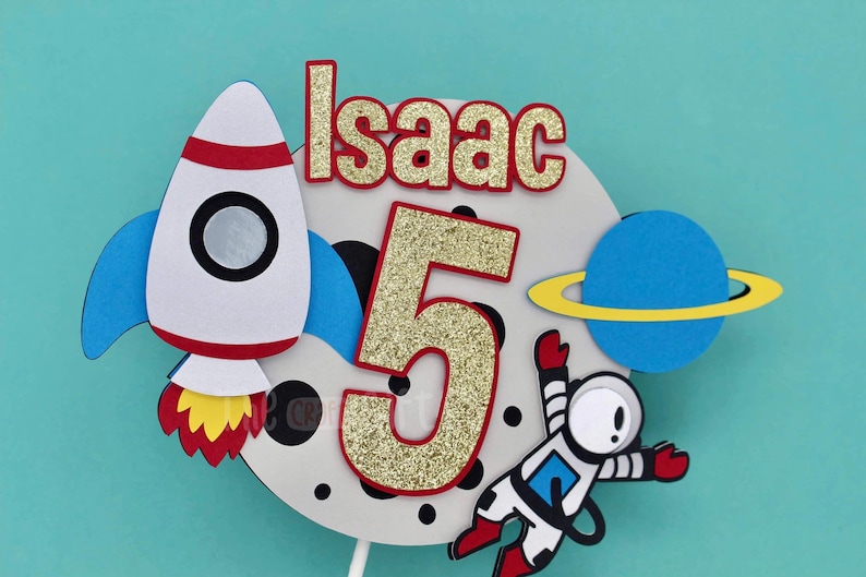 Puede incluir: Un adorno para pastel blanco con un cohete, un astronauta, un planeta y el n&uacute;mero 5 en purpurina dorada. El texto "Isaac" est&aacute; escrito en letras rojas.