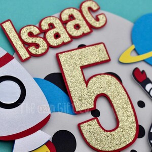 Puede incluir: N&uacute;mero cinco de purpurina dorada con contorno rojo, un cohete blanco con ribetes rojos, un planeta azul con un anillo amarillo y un astronauta blanco con un traje azul y un casco gris. El nombre "Isaac" est&aacute; escrito en letras doradas de purpurina con contorno rojo.