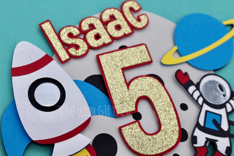 Puede incluir: N&uacute;mero cinco de purpurina dorada con contorno rojo, un cohete blanco con ribetes rojos, un planeta azul con un anillo amarillo y un astronauta blanco con un traje azul y un casco gris. El nombre "Isaac" est&aacute; escrito en letras doradas de purpurina con contorno rojo.