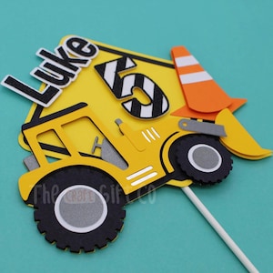 May include: Yellow construction vehicle cake topper with a black and white striped number 5 and the name Luke on a yellow construction sign. The topper also includes an orange traffic cone.