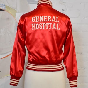 May include: A red satin jacket with the words "GENERAL HOSPITAL" embroidered in white on the back. The jacket has a ribbed collar, cuffs, and hem with white and red stripes. The jacket is displayed on a mannequin.