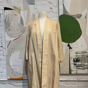 May include: A beige kimono-style robe with a shawl collar and three-quarter sleeves. The robe is displayed on a mannequin against a backdrop of abstract art and a white brick wall. The robe has a subtle textured pattern.