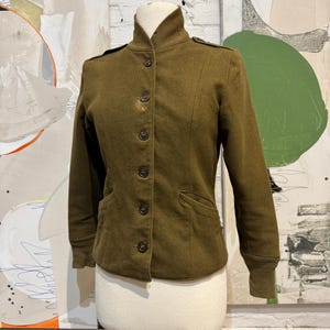 Op de afbeelding: Olijfgroene jas met een hoge kraag en knoopsluiting. De jas heeft twee voorzakken en schouderkleppen. De mouwen hebben geribde manchetten. De jas wordt op een mannequin gepresenteerd.
