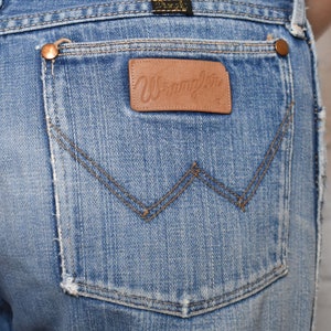 K&ouml;nnte beinhalten: Eine blaue Jeanshose mit einem braunen Leder-Patch auf der Ges&auml;&szlig;tasche, auf dem "Wrangler" steht.