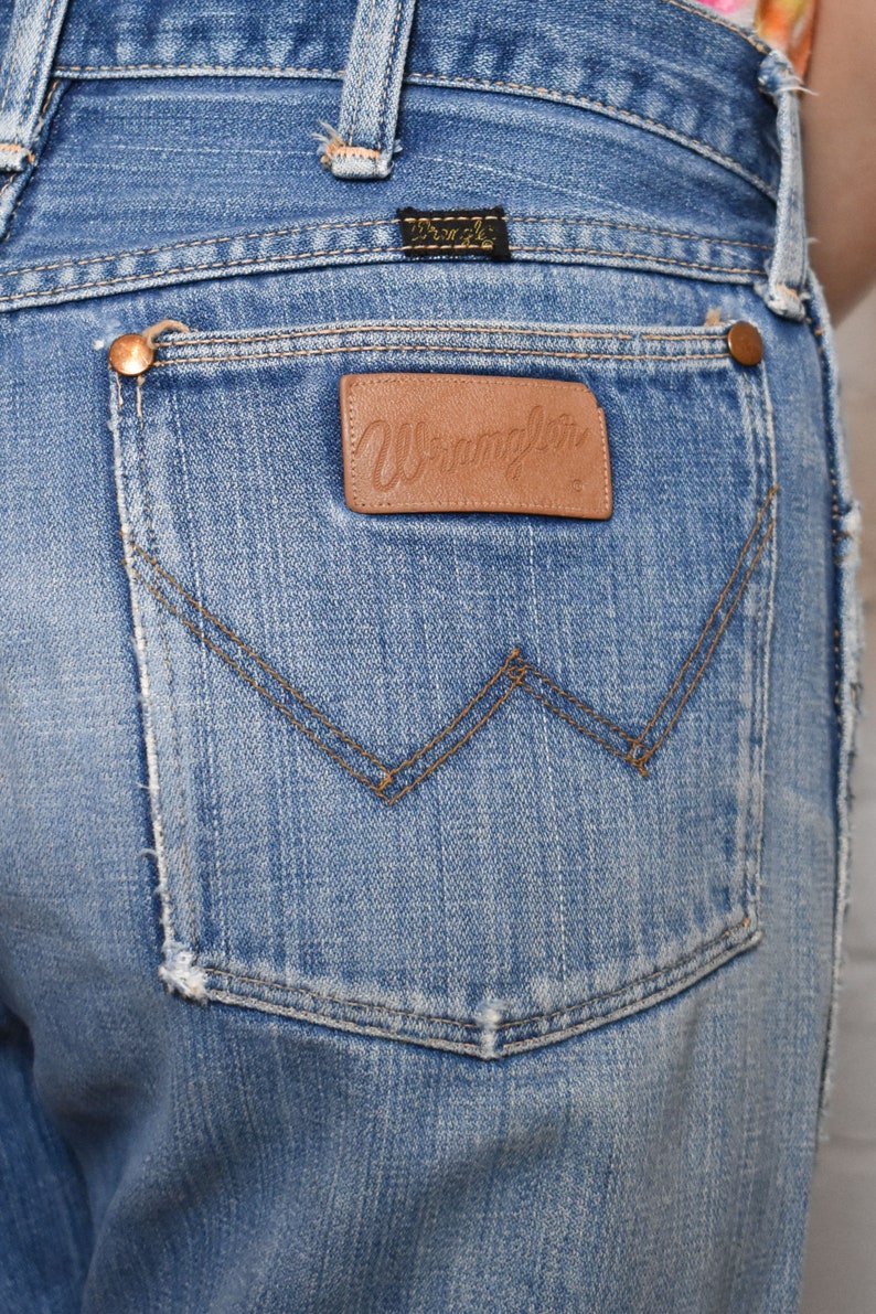 K&ouml;nnte beinhalten: Eine blaue Jeanshose mit einem braunen Leder-Patch auf der Ges&auml;&szlig;tasche, auf dem "Wrangler" steht.
