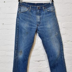 May include: Faded blue denim jeans hanging on a black hanger against a white brick wall. The jeans show signs of wear, including discoloration and a small hole. The classic five-pocket design is visible.