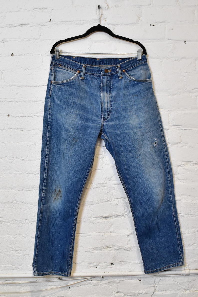 May include: Faded blue denim jeans hanging on a black hanger against a white brick wall. The jeans show signs of wear, including discoloration and a small hole. The classic five-pocket design is visible.