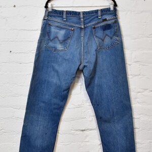 May include: Blue denim jeans hanging on a black hanger. The jeans have a classic five-pocket design, with the signature 'W' stitching on the back pockets. The denim has a faded look, with some wear and tear visible.