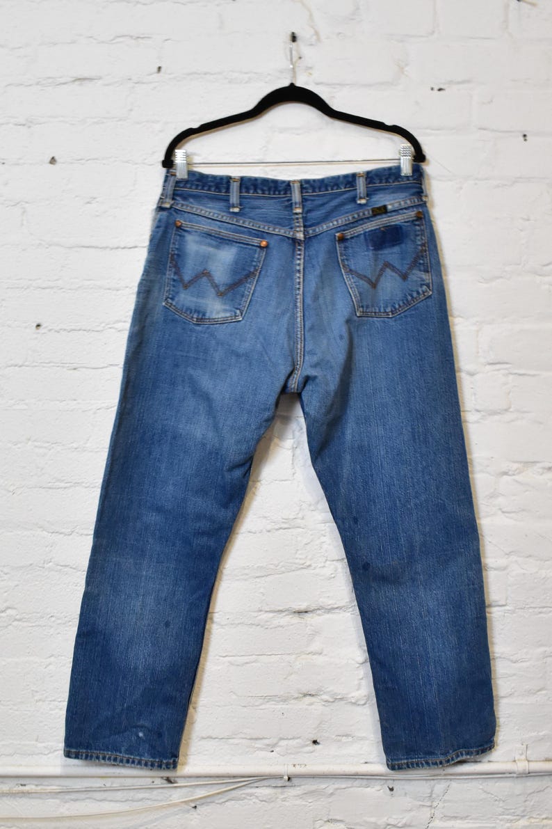 May include: Blue denim jeans hanging on a black hanger. The jeans have a classic five-pocket design, with the signature 'W' stitching on the back pockets. The denim has a faded look, with some wear and tear visible.