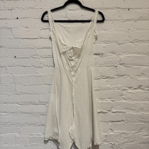 May include: A white vintage slip with a square neckline and delicate lace trim. The slip features a button-up front and a flared skirt, hanging on a black hanger against a white brick wall.