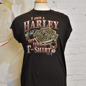 May include: Black Harley-Davidson t-shirt featuring the text "I Own A Harley, Not Just A T-Shirt" and the Harley-Davidson logo in silver, gold, and orange. The t-shirt is displayed on a mannequin.