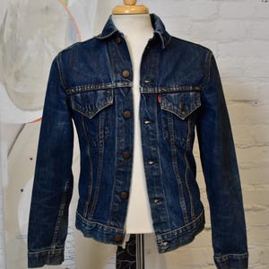 May include: A dark blue denim jacket with button fastenings, two chest pockets, and a small red label. The jacket is displayed on a mannequin, highlighting its vintage style and robust fabric. A timeless wardrobe staple.