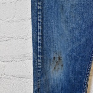 May include: Close-up of a pair of blue denim jeans with visible wear and tear. The jeans have a faded appearance with dark stains on the leg. The stitching is visible along the seams and the edge of the jeans.