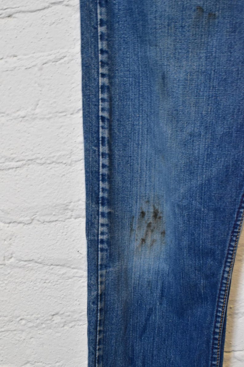 May include: Close-up of a pair of blue denim jeans with visible wear and tear. The jeans have a faded appearance with dark stains on the leg. The stitching is visible along the seams and the edge of the jeans.