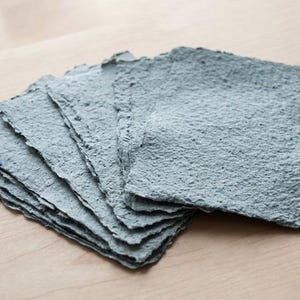 May include: A stack of six square sheets of handmade paper in a muted blue-gray color. Each sheet features a thick, fibrous texture and uneven, deckled edges, creating a layered appearance with visible variations in the paper surface density.