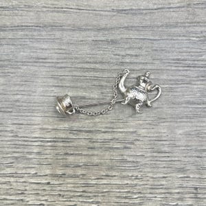 May include: A silver-toned brooch featuring a teapot design, connected by a chain to a decorative pin. The teapot has a detailed handle, spout, and lid. The pin has a decorative design. The brooch is set against a gray wooden surface.