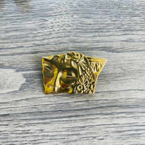 May include: A gold-toned metal brooch with an intricate design featuring a face and floral details.