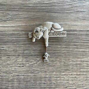 May include: A silver-toned cat figurine hanging from a decorative ledge, dangling a small silver-toned mouse figurine from its paw.
