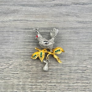May include: A silver and gold tone brooch featuring a bird perched on a branch with green rhinestones. The bird has a red rhinestone eye and a pear-shaped dangle.
